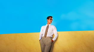 Model wearing a white shirt, textured neutral tie and taupe trousers against a yellow wall and blue sky